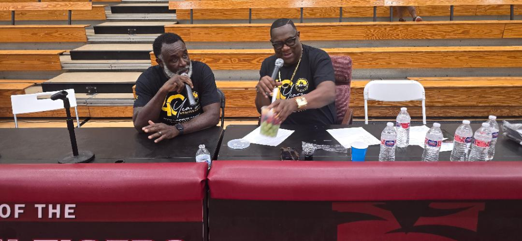 Two AMB leaders in 40th anniversary shirts speak at a panel table inside Forbes Arena with bleachers in the background