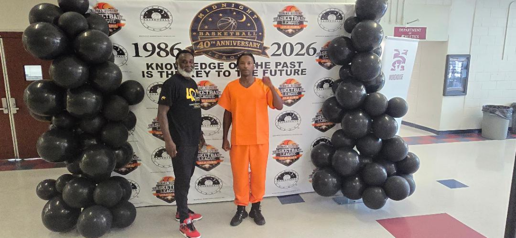Two men pose at the 40th Anniversary AMB step-and-repeat — one in black AMB anniversary shirt, one in bright orange outfit