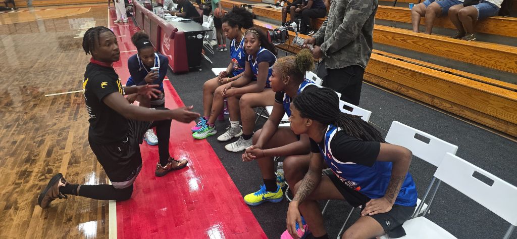 Coach in black addresses women's basketball team in blue jerseys seated on the bench at Forbes Arena — Coca-Cola scorer's table in background